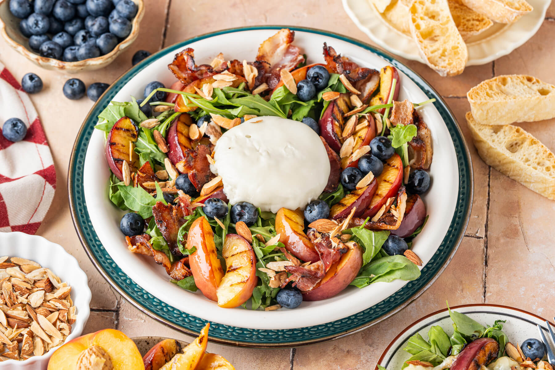 A salad bowl containing grilled peaches, toasted almonds, blueberries, crumbled bacon, and burrata cheese on a bed of green arugula drizzled with balsamic salad dressing.