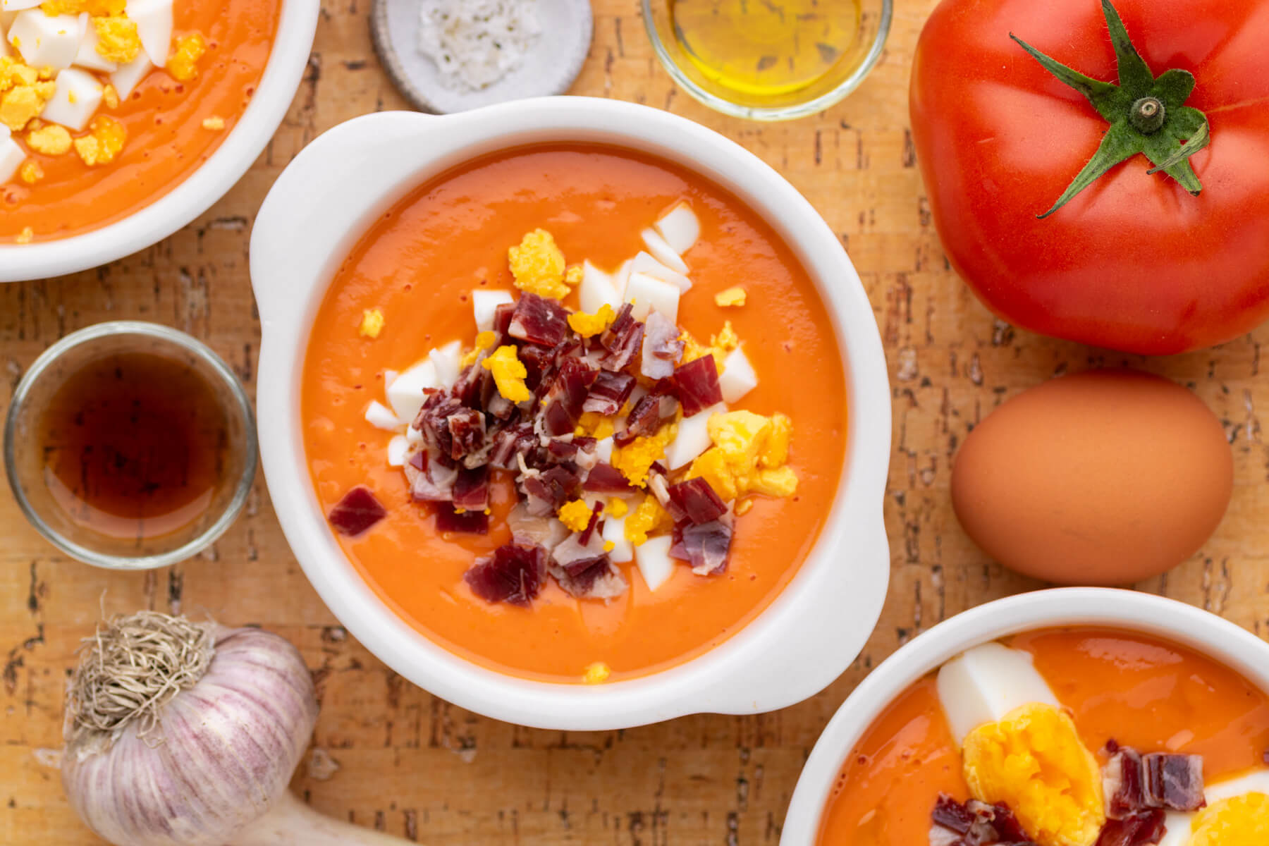 Three white bowls of vibrant orange soup garnished with chopped hard boiled eggs and jamón ibérico surrounded by recipe ingredients.