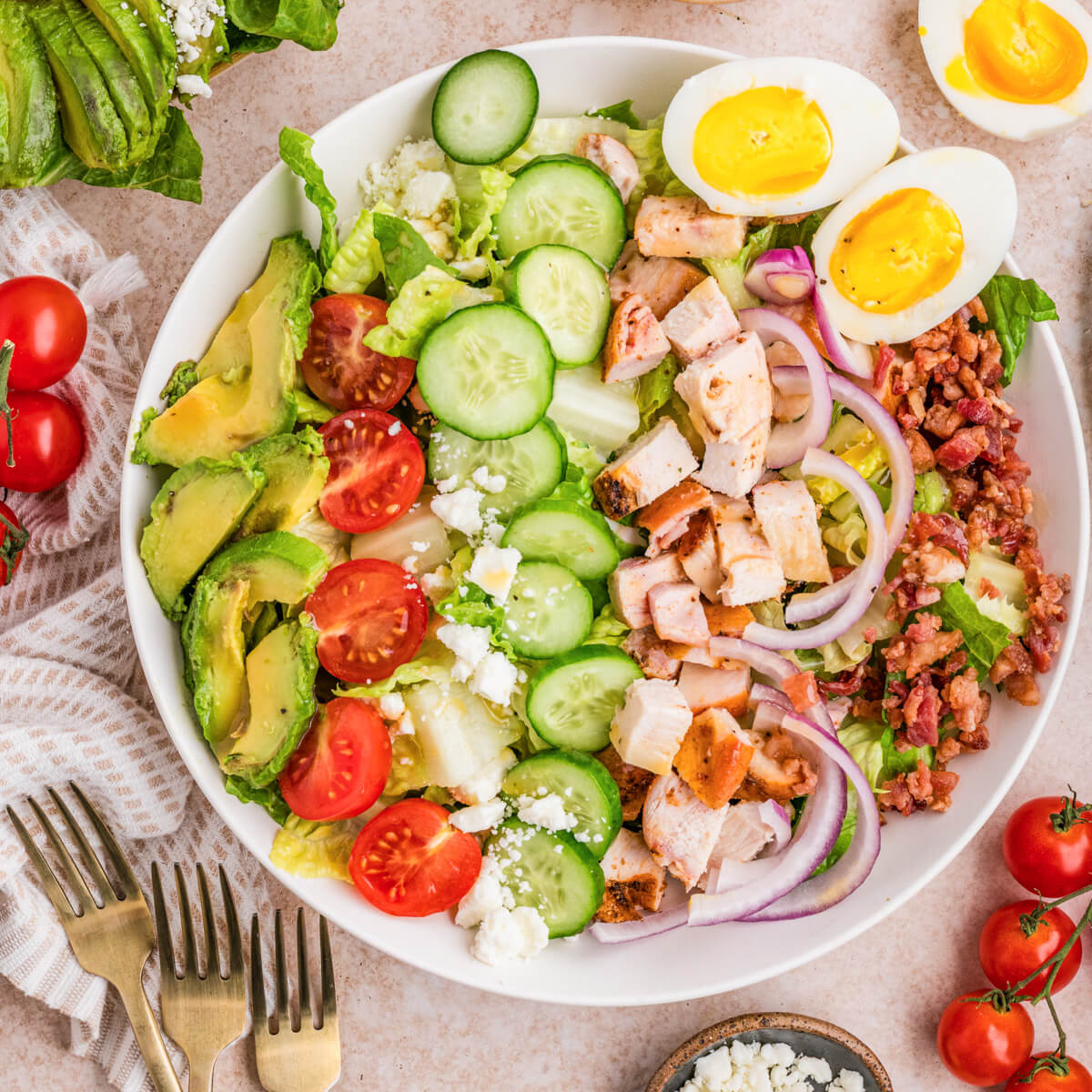 A white salad bowl containing a tastefully arranged Chicken Cobb Salad with grape tomatoes, boiled eggs, bacon, avocado, red onions, sliced chicken, and cucumbers.
