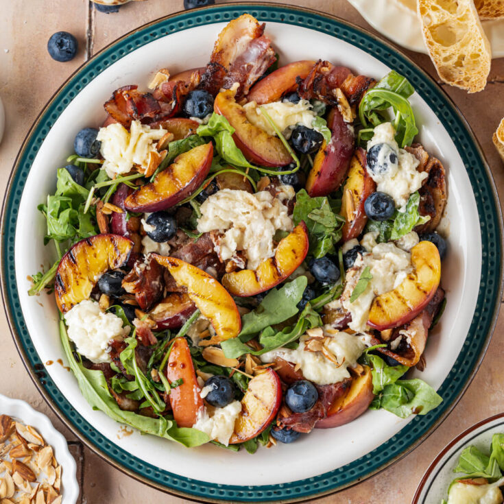 A salad bowl containing grilled peaches, toasted almonds, blueberries, crumbled bacon, and burrata cheese on a bed of green arugula drizzled with balsamic salad dressing.