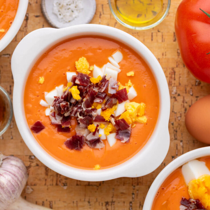 A white bowl of vibrant orange soup garnished with chopped hard boiled eggs and jamón ibérico.