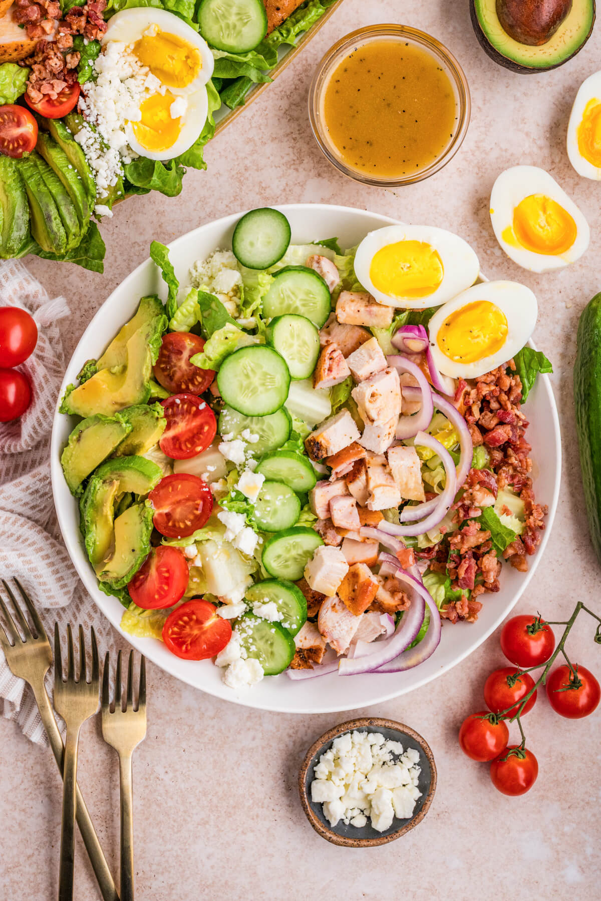 A white salad bowl containing a tastefully arranged Chicken Cobb Salad with grape tomatoes, boiled eggs, bacon, avocado, red onions, sliced chicken, and cucumbers surrounded by ingredients.