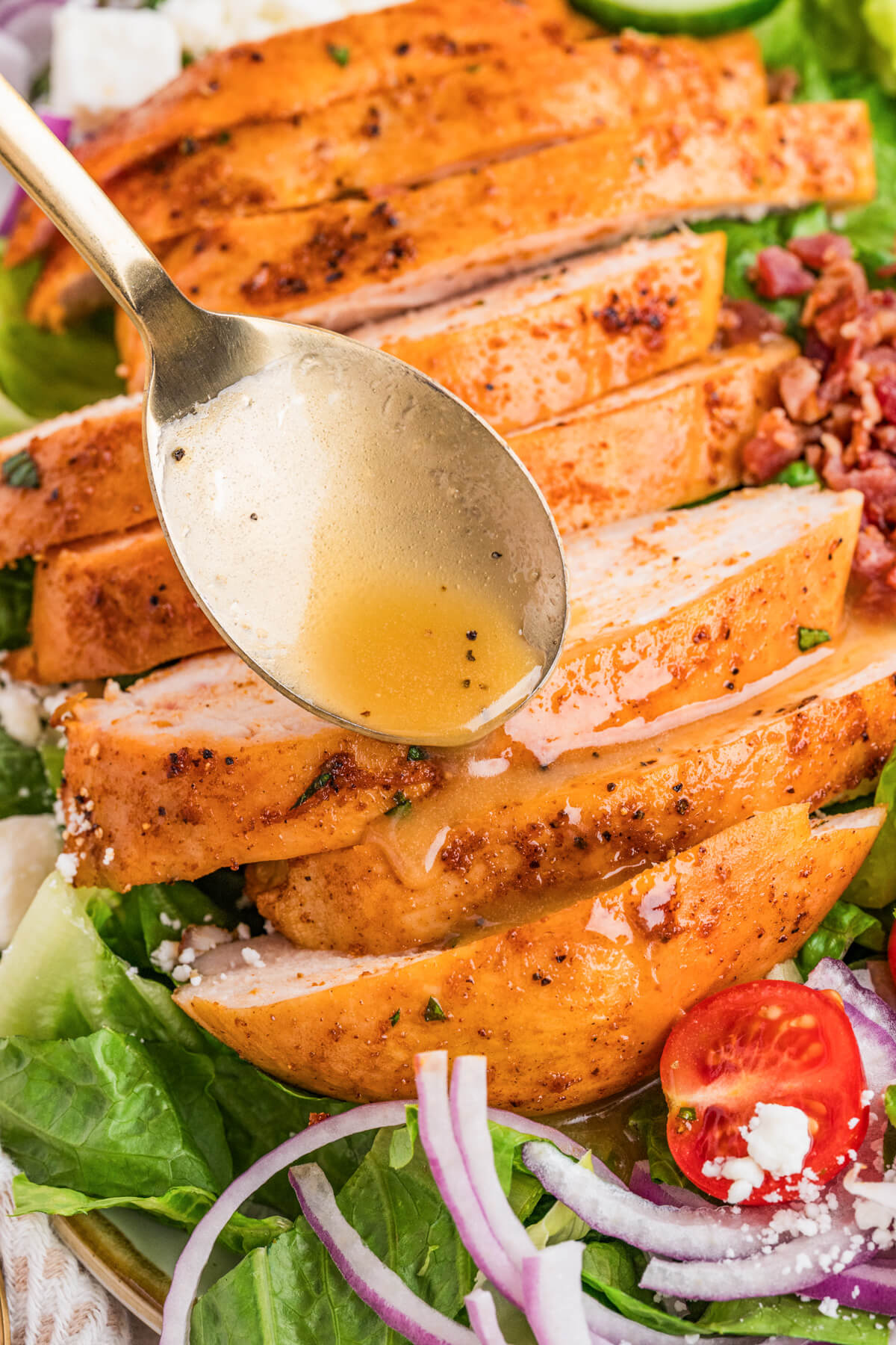 A spoon drizzles salad dressing over a sliced smoked chicken breast on a cobb salad.