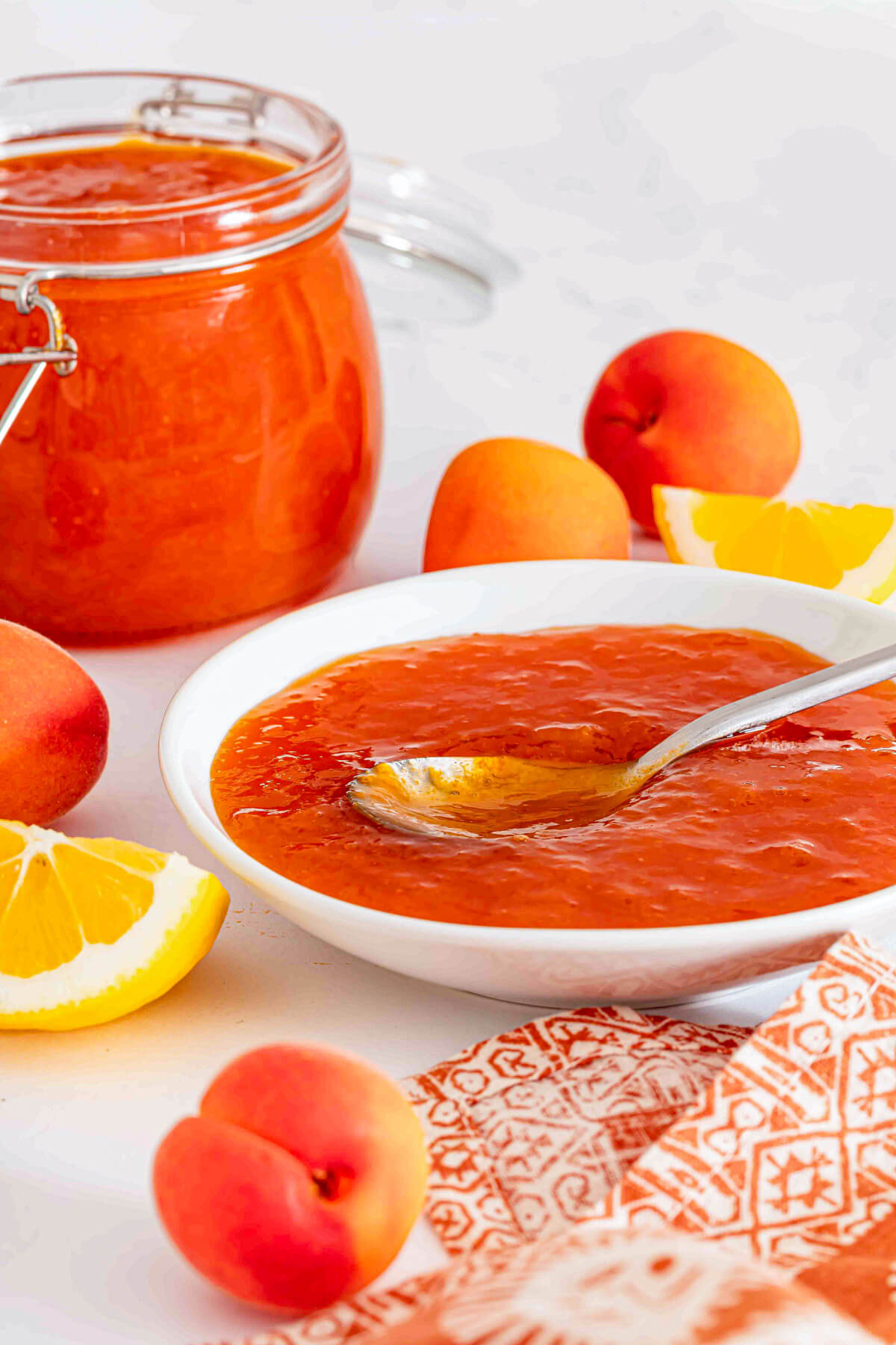 Apricot jam in a small white bowl surrounded by fresh apricots, lemons, and a jar of the same jam.