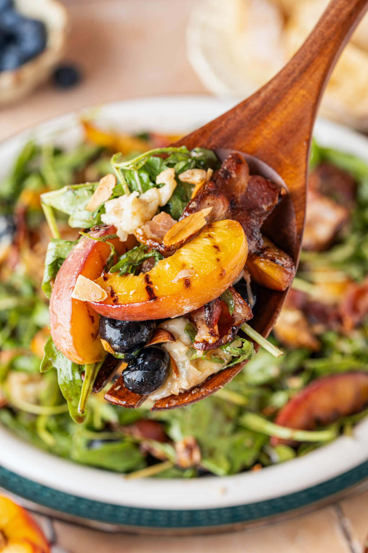 A wooden salad server containing baby arugula, blueberries, a grilled peach slice, almonds, bacon, and burrata cheese.