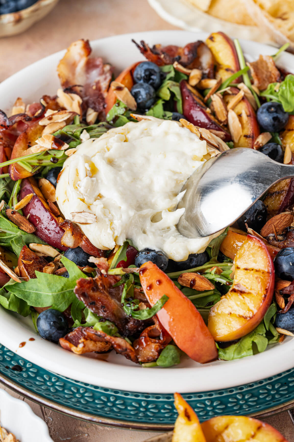 A spoon spreads a ball of creamy burrata cheese over a grilled peach salad.