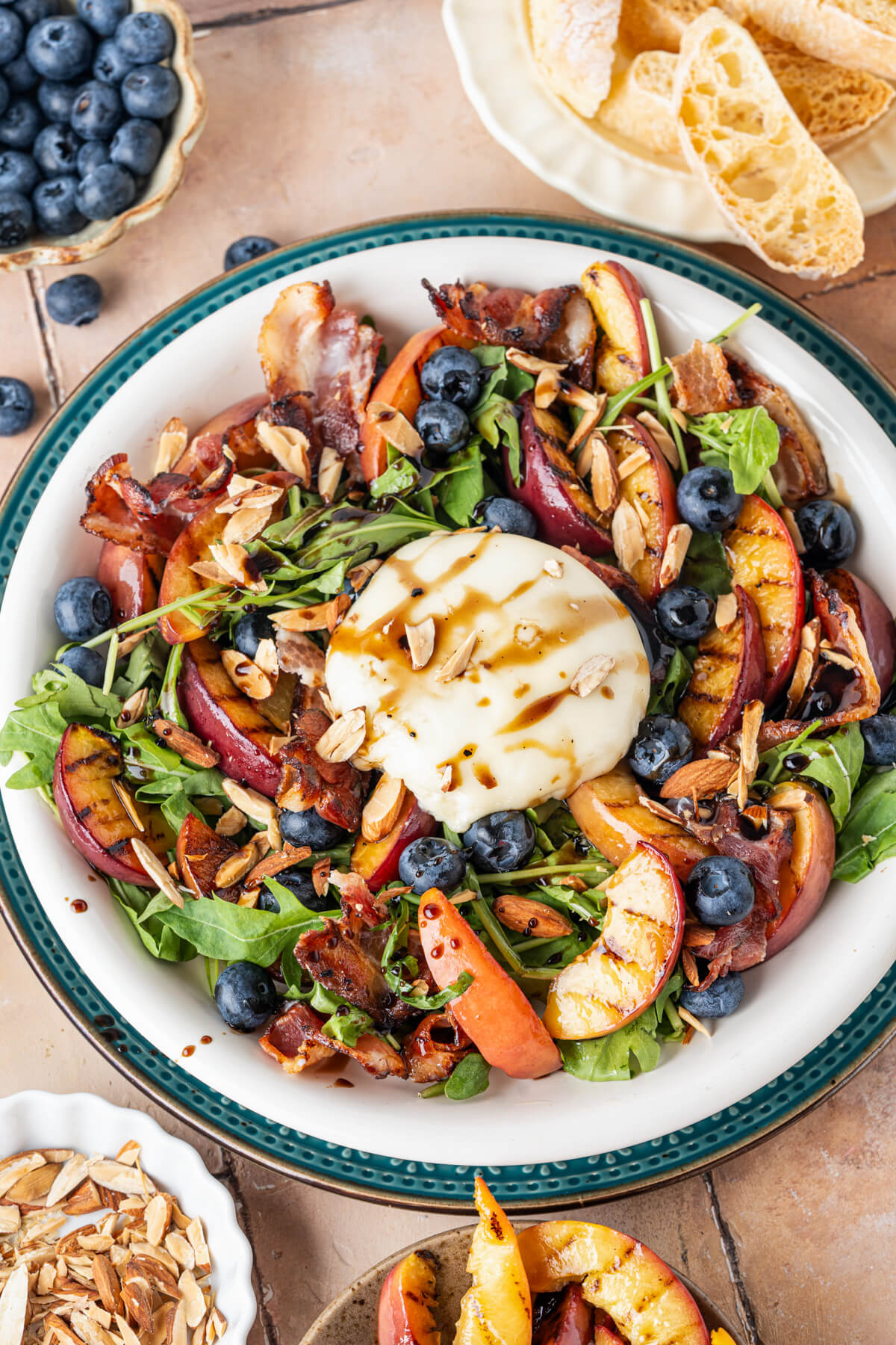 A salad bowl containing grilled peaches, toasted almonds, blueberries, crumbled bacon, and burrata cheese on a bed of green arugula drizzled with balsamic salad dressing.