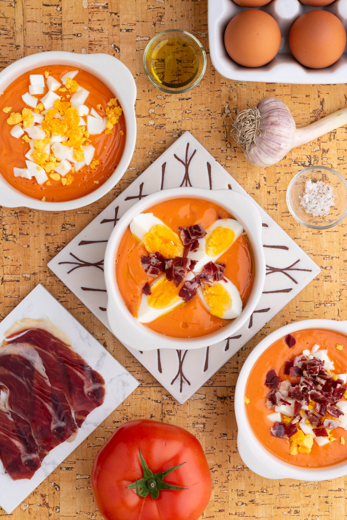 Three white bowls of vibrant orange soup garnished with chopped hard boiled eggs and jamón ibérico surrounded by recipe ingredients.