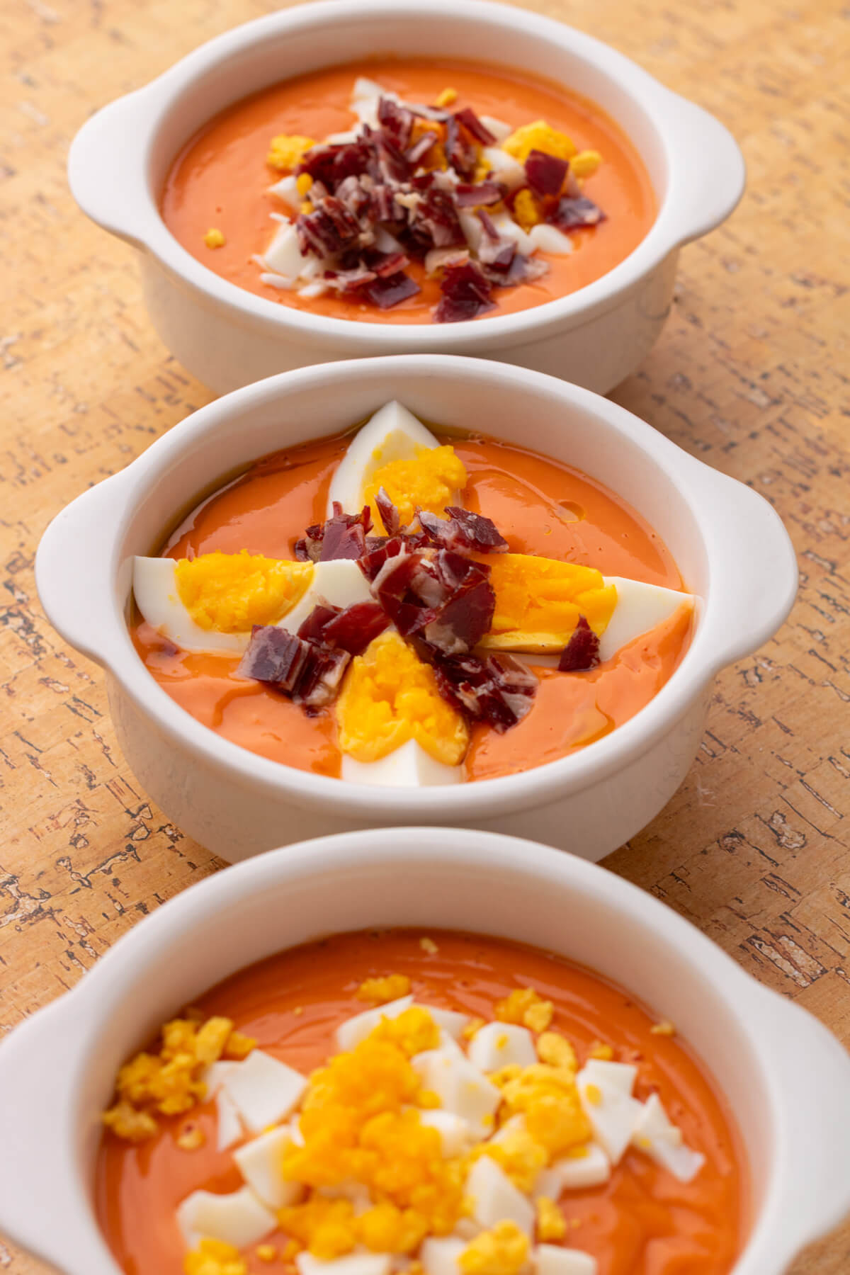 Three white bowls of vibrant orange soup garnished with chopped hard boiled eggs and jamón ibérico.