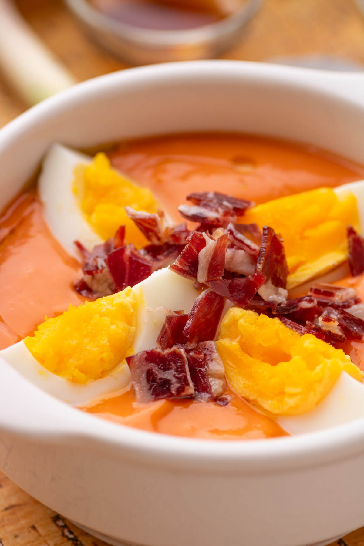 A white soup bowl filled with vibrant orange Salmorejo garished with sliced egg quarters and diced Iberico jamon.