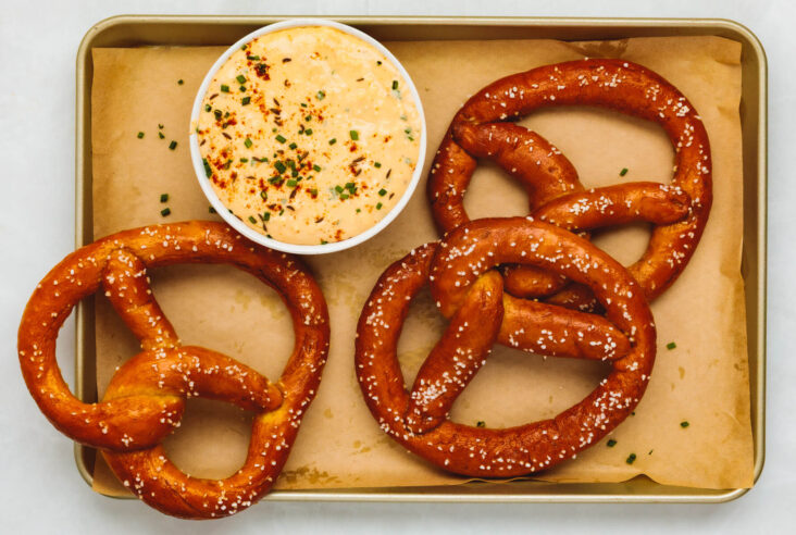 A tray of deep brown homemade pretzels topped with coarse salt beside a small bowl of Obatzda dip.