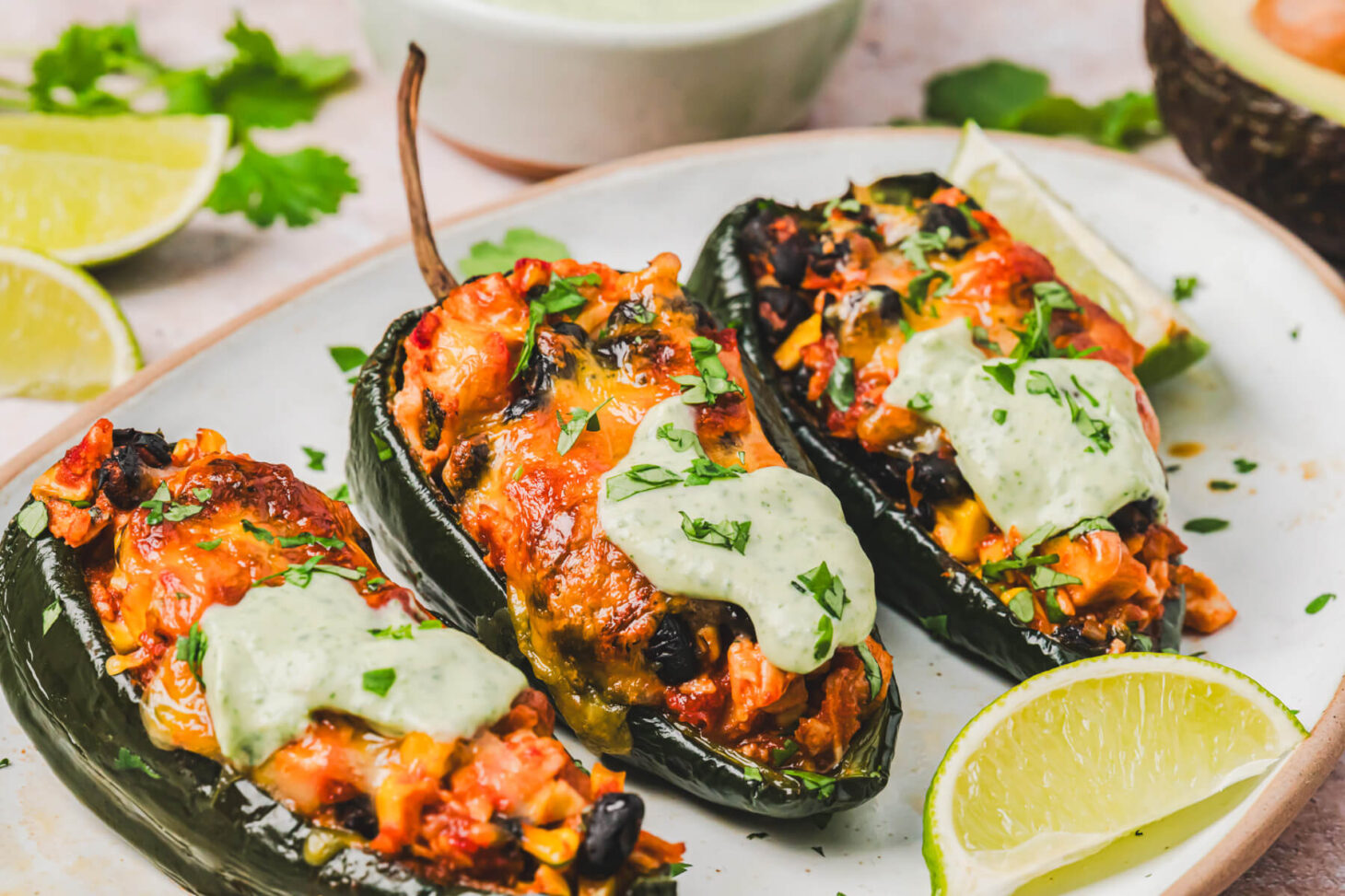 Three stuffed poblano peppers topped with a layer of melted cheese and a dollop of cilantro crema on a plate.