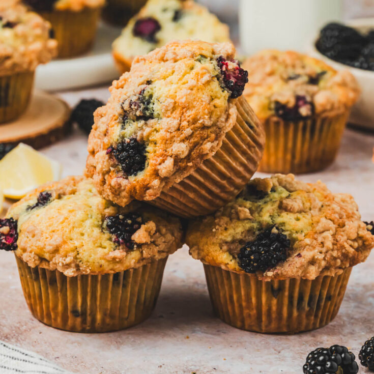 Three golden baked blackberry muffins stacked high in front of more muffins and fresh blackberries.
