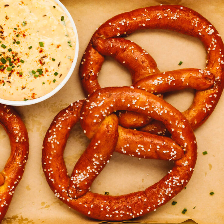 Two deep brown homemade pretzels topped with coarse salt beside a small bowl of obatzda dip.
