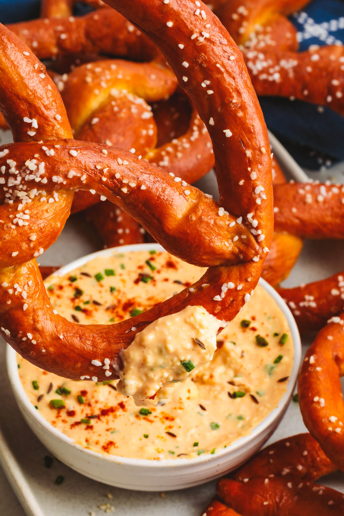 A glossy, salty, deep brown homemade pretzel being dipped into a bowl of Obatzda cheese dip.