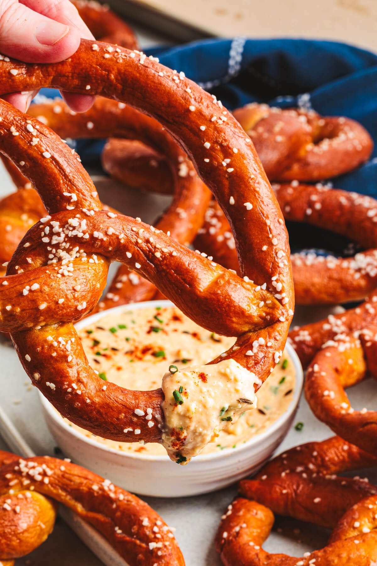 A hand dips a glossy, salty, deep brown homemade pretzel being dipped into a bowl of Obatzda cheese dip.