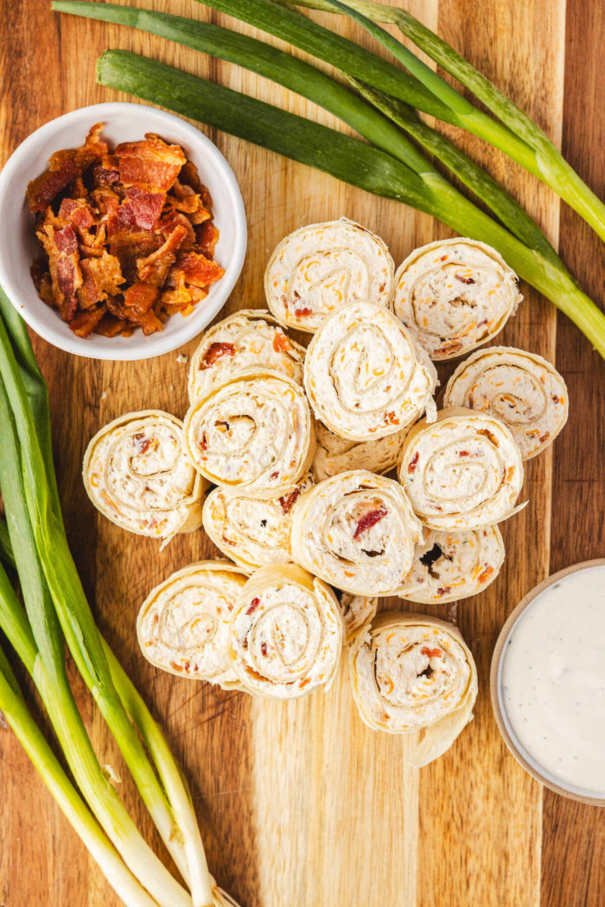 A pile of tortilla pinwheels filled with cream cheese mixture containing bacon, cheddar cheese, and shredded chicken beside green onions on a wooden serving board.