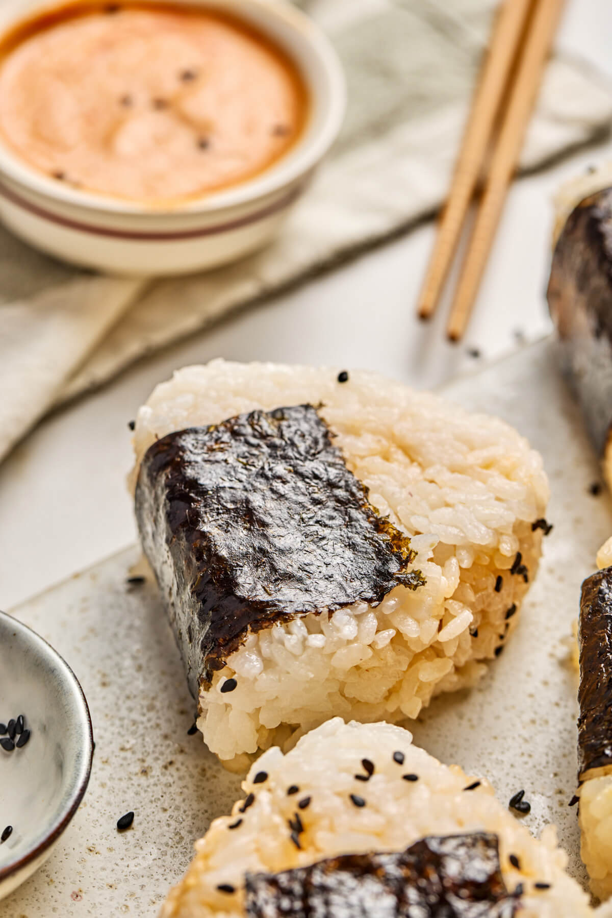 One Japanese Onigiri wrapped in nori and garnished with black sesame seeds beside a small bowl of sriracha mayonnaise sauce.