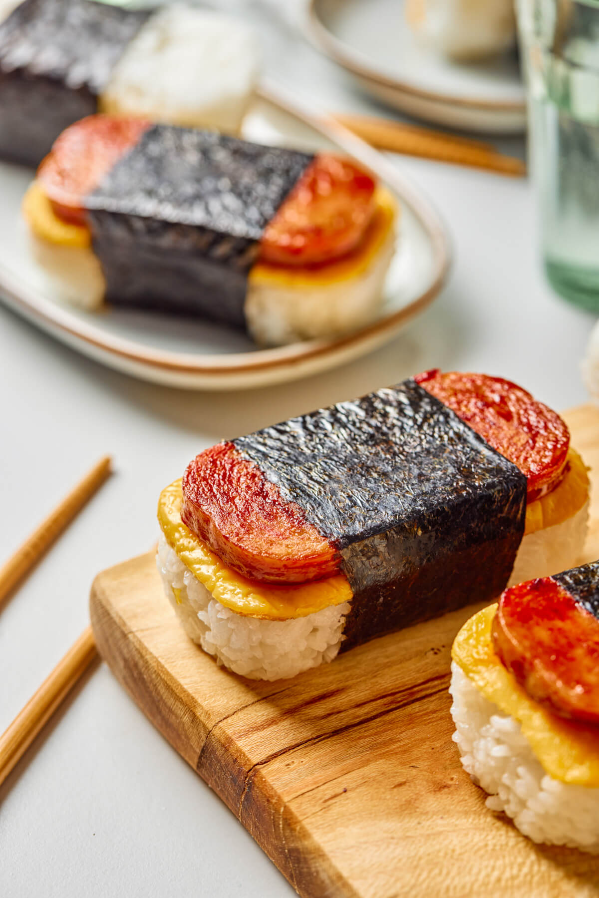 Two Spam and Egg Musubi wrapped in nori strips on a wooden serving board and on a plate.