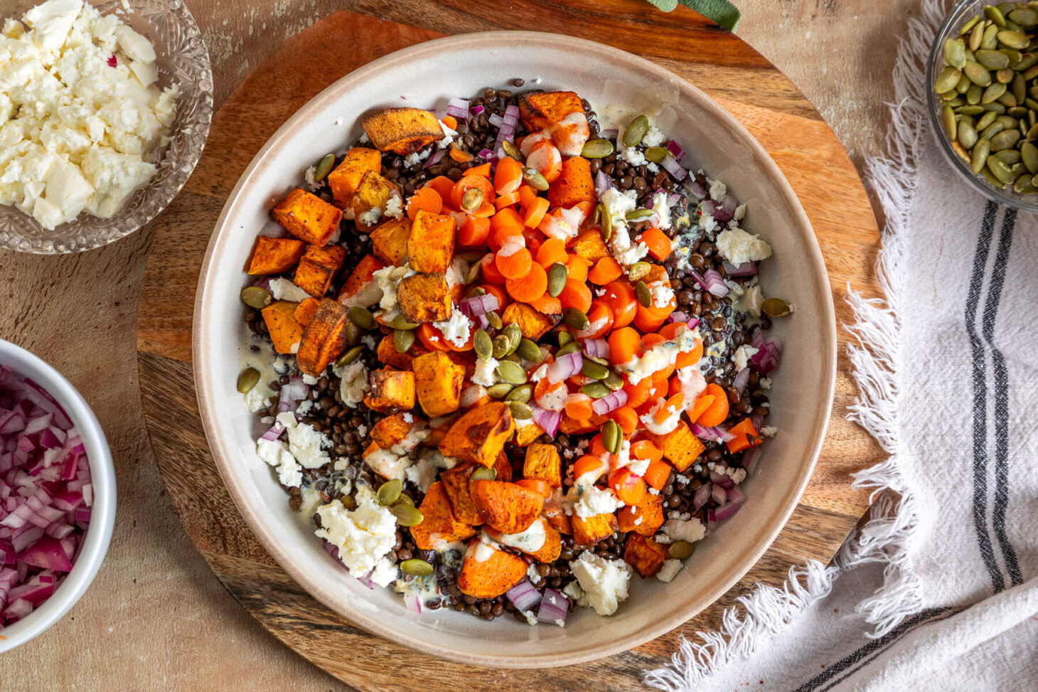 A vibrant orange Sweet Potato Lentil Bowl topped with feta cheese, and toasted pepitas on a dinner table.