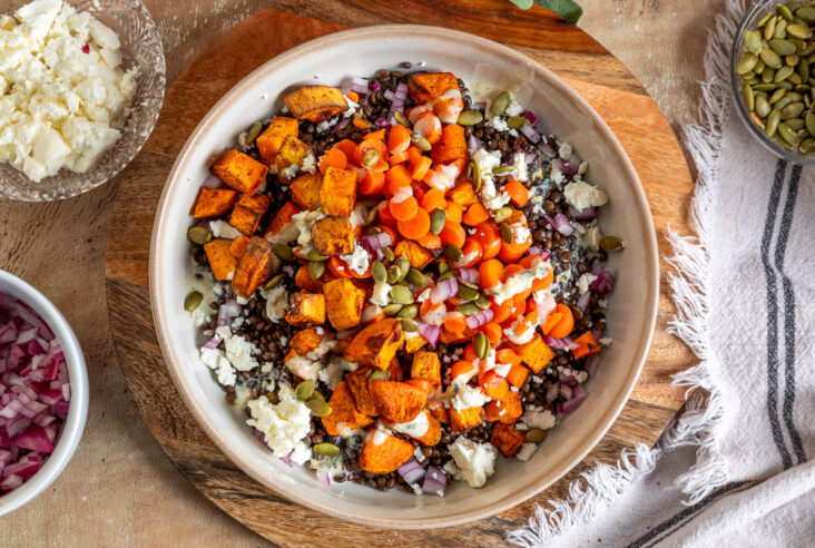 A vibrant orange Sweet Potato Lentil Bowl topped with feta cheese, and toasted pepitas on a dinner table.