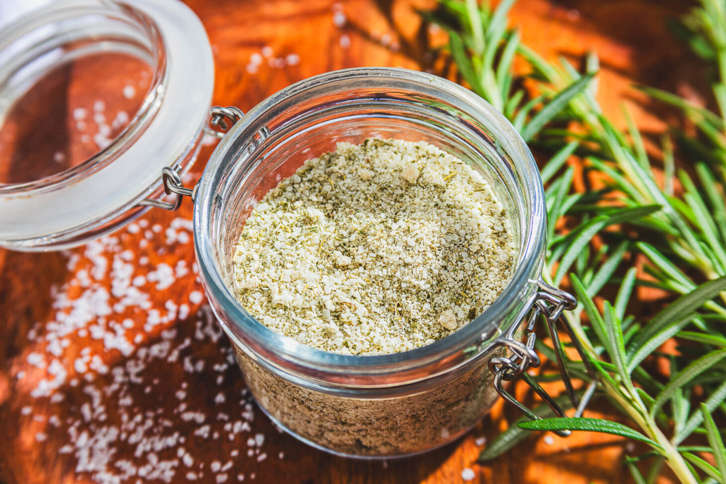a glass jar of rosemary salt surrounded by fresh rosemary on a wooden platter.