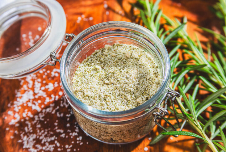 a glass jar of rosemary salt surrounded by fresh rosemary on a wooden platter.