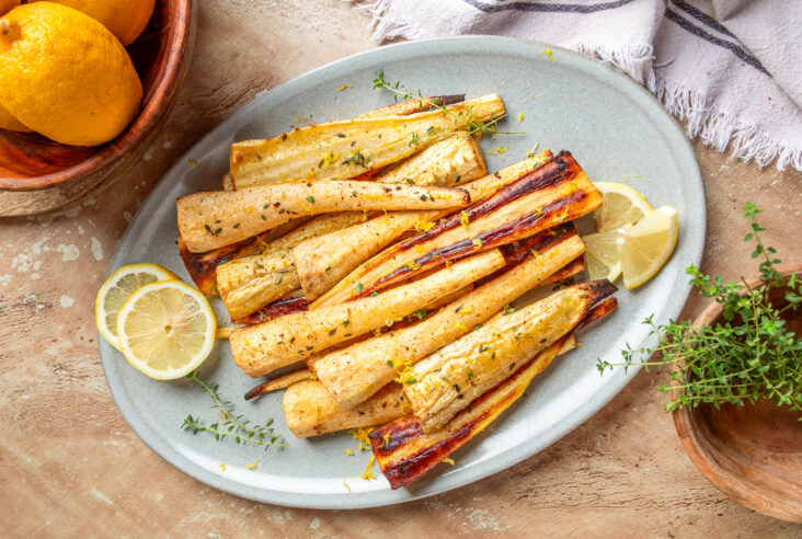 A light grey platter holds golden caramelized roasted parsnips garnished with fresh green herbs and lemon zest.