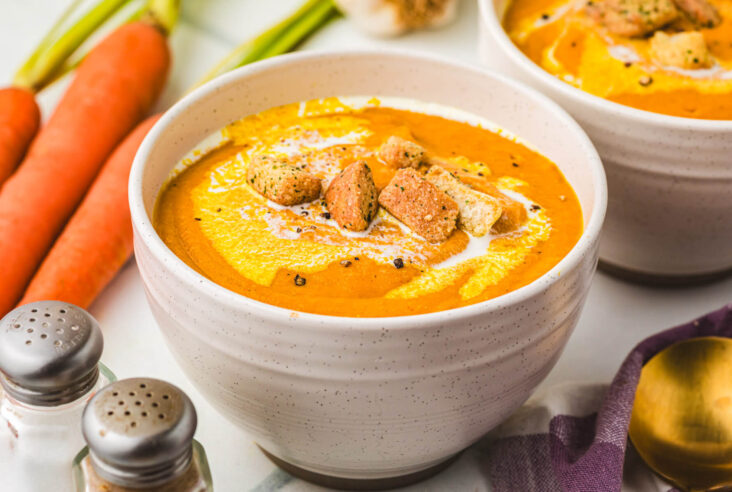 Two soup bowls of vibrant Carrot Lentil Soup garnished with a swirl of sour cream and toasted croutons.