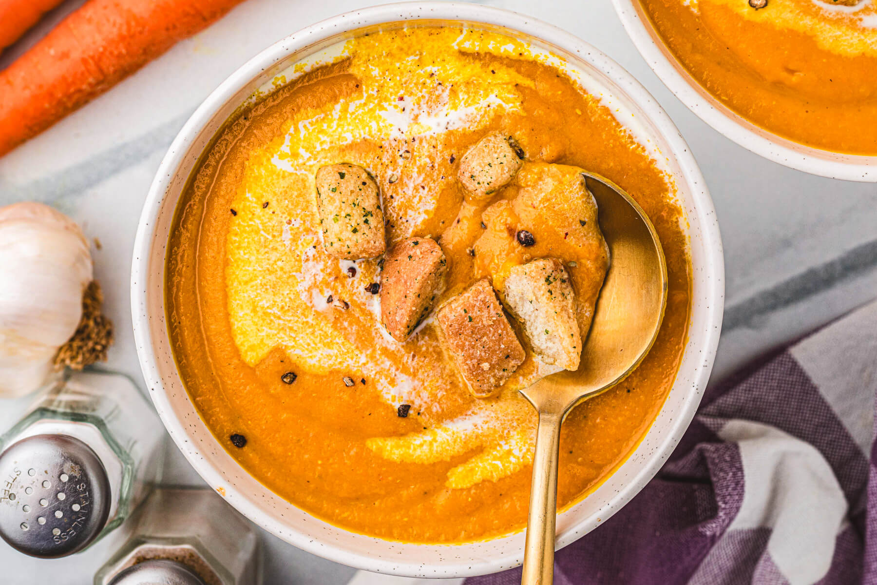 A spoon in a white soup bowl filled with vibrant orange Carrot Lentil Soup garnished with a swirl of sour cream and toasted croutons.