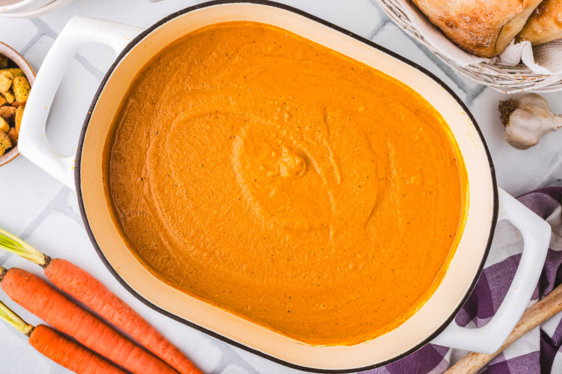 A large oval soup pot filled vibrant orange carrot lentil soup surrounded by soup ingredients.