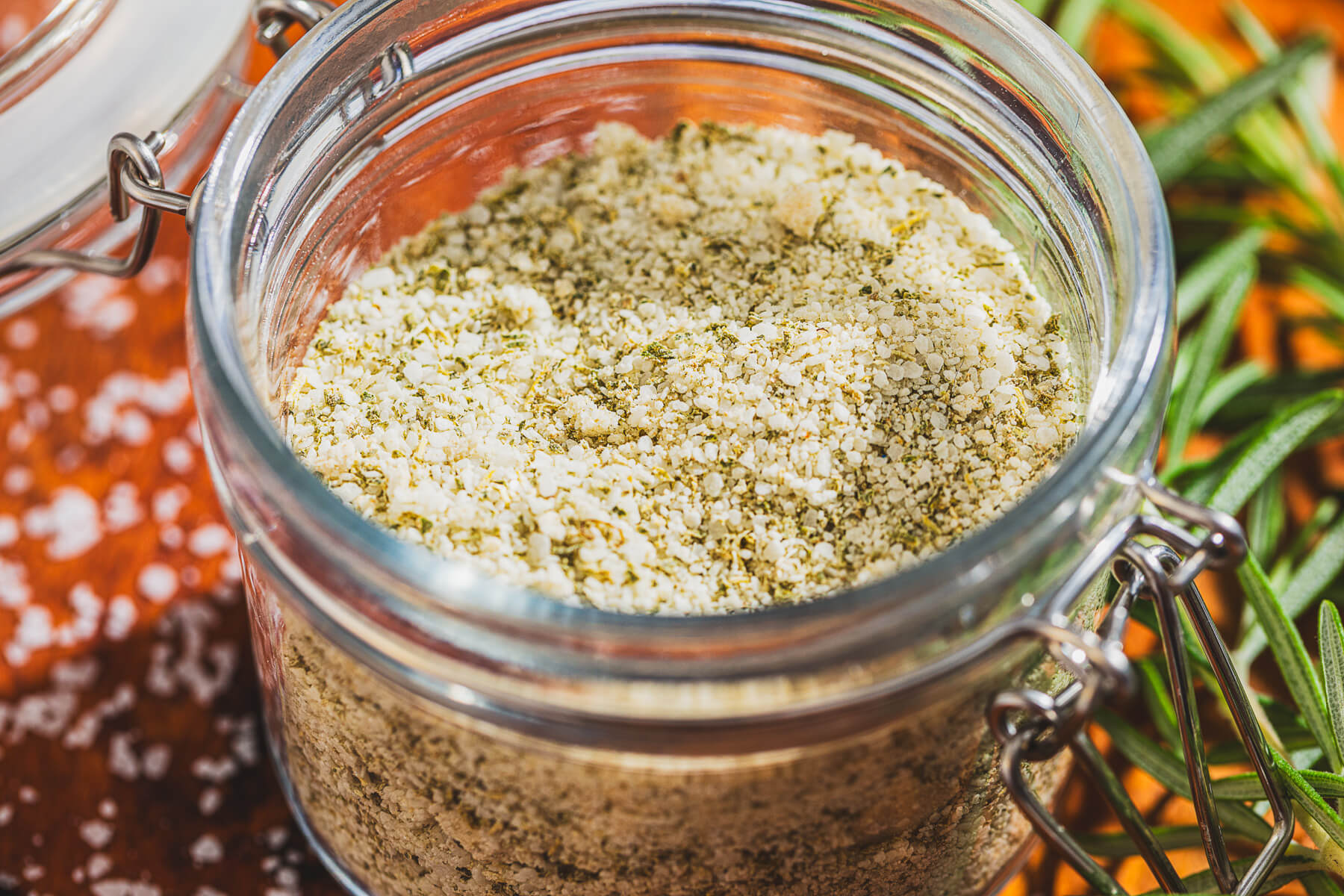 a glass jar of rosemary salt surrounded by fresh rosemary on a wooden platter.