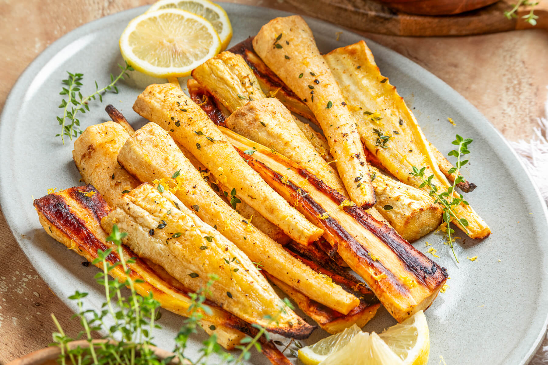 A light grey platter holds golden caramelized roasted parsnips garnished with fresh green herbs and lemon zest.