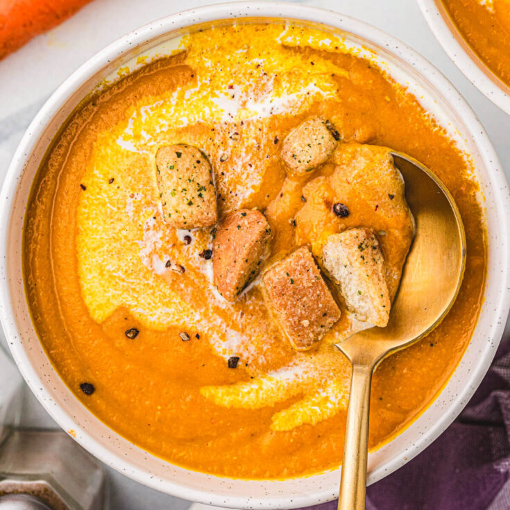 A soup bowl full of vibrant Carrot Lentil Soup garnished with a swirl of sour cream and toasted croutons.
