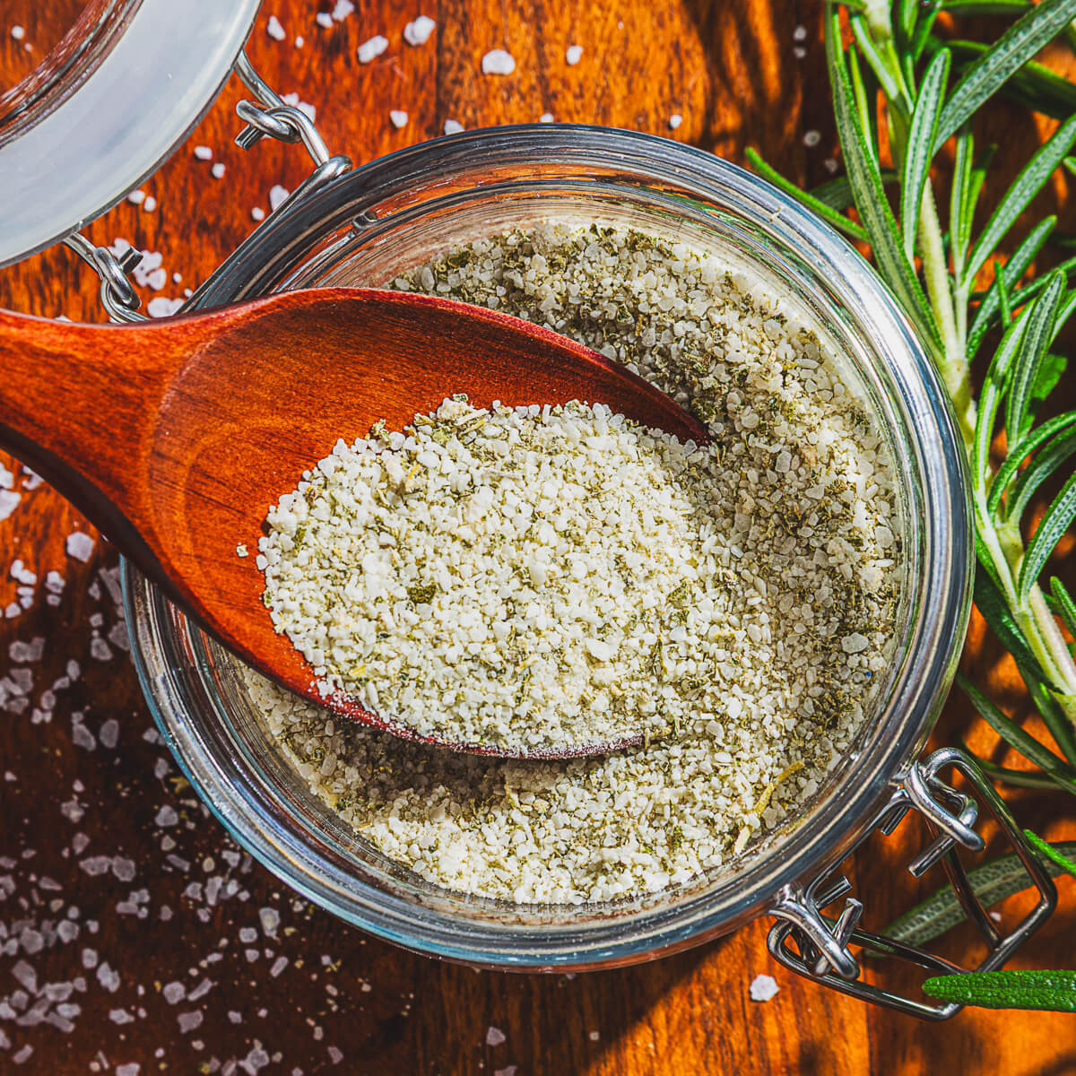 A glass jar of rosemary salt and a wooden spoon containing the same salt surrounded by fresh rosemary.