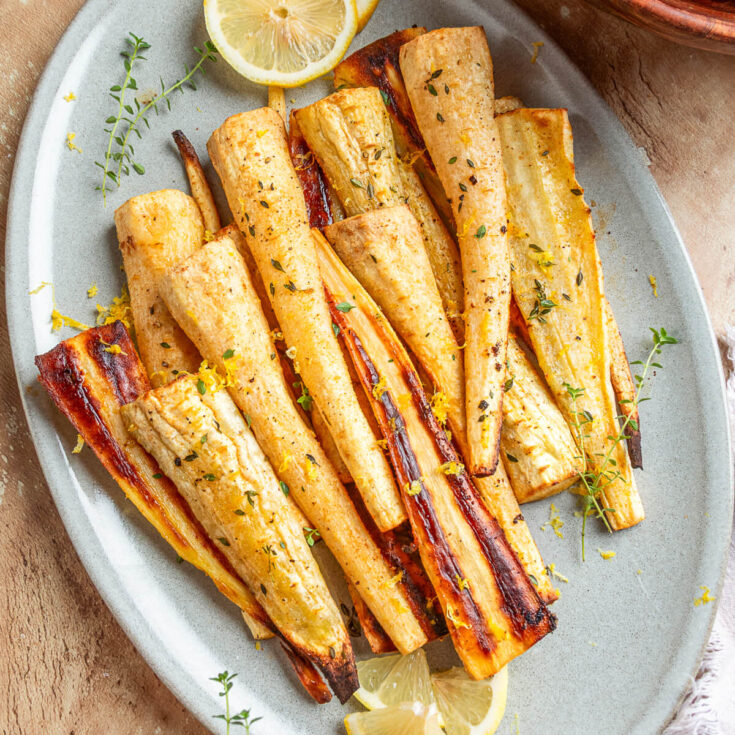 A light grey platter holds golden caramelized roasted parsnips garnished with fresh green herbs and lemon zest.