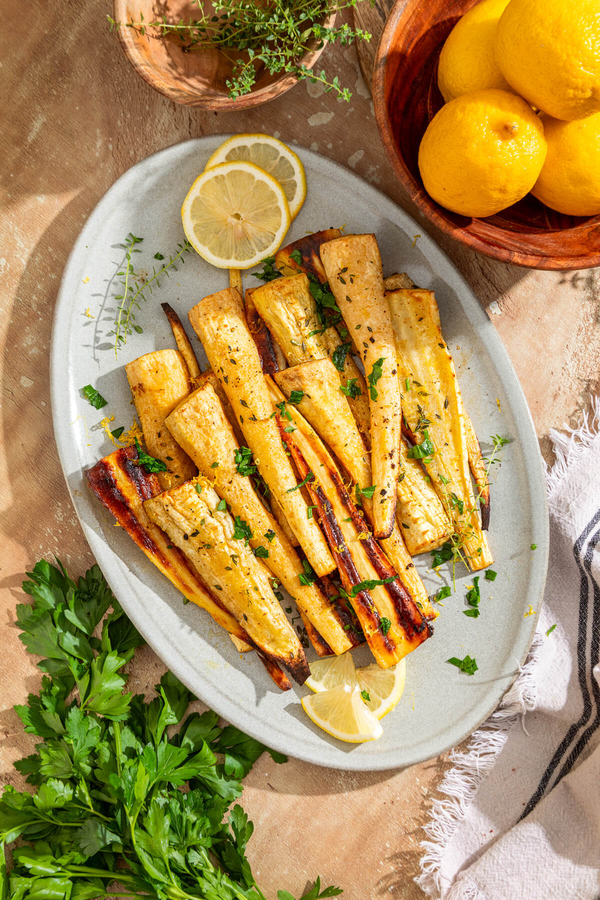 A light grey platter holds golden caramelized roasted parsnips garnished with fresh green herbs and lemon zest.