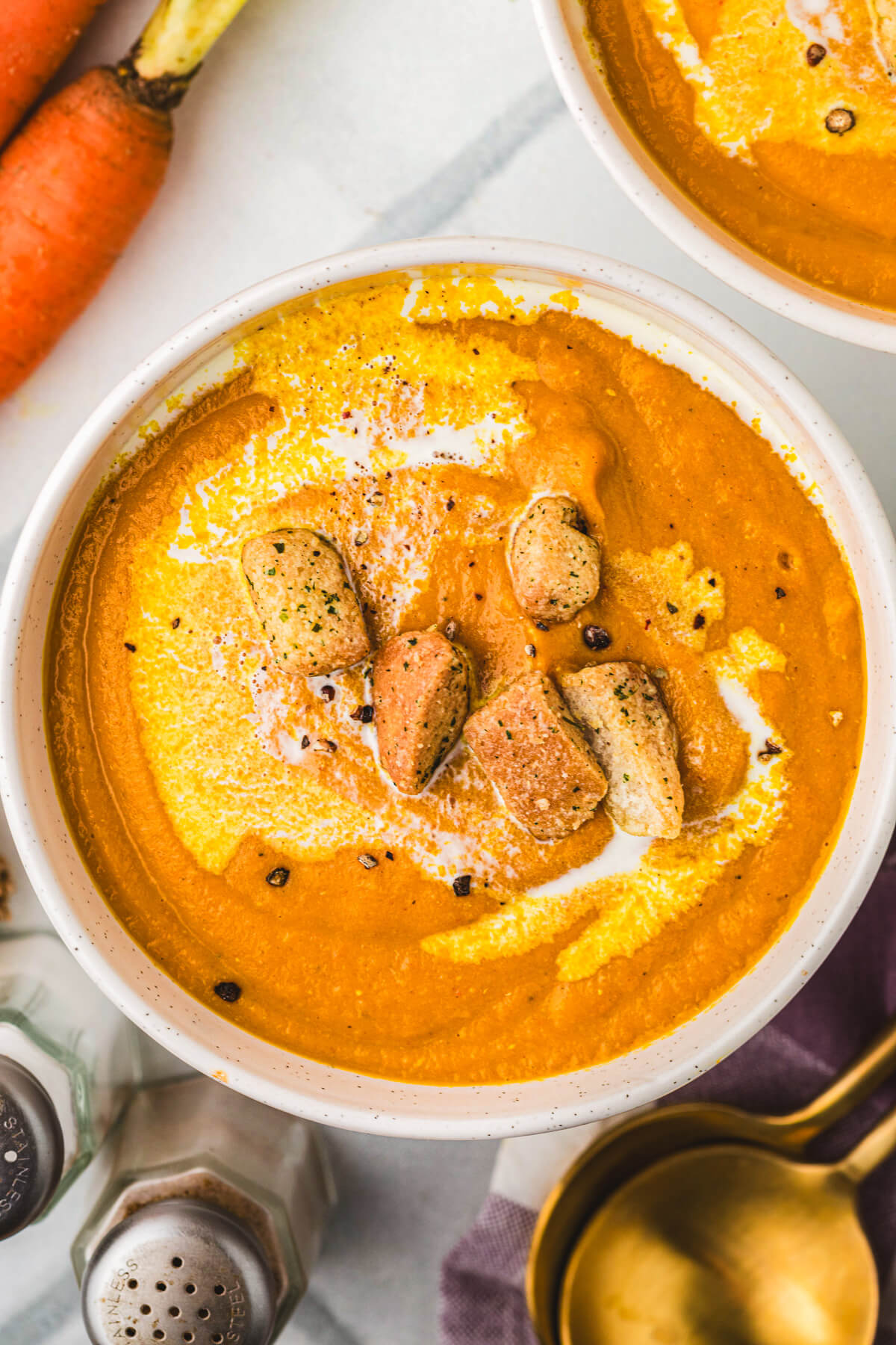 A white soup bowl filled with vibrant orange Carrot Lentil Soup garnished with a swirl of sour cream and toasted croutons.
