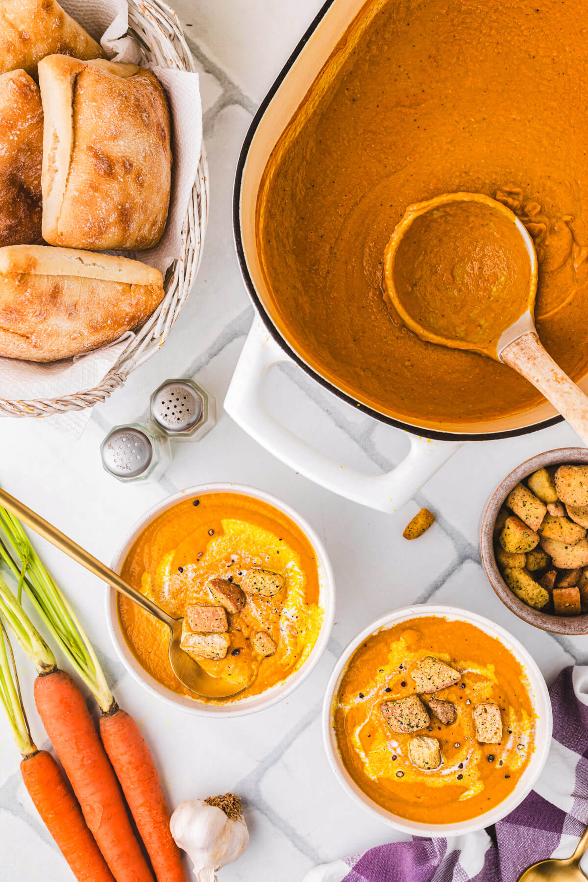 Two soup bowls of vibrant Carrot Lentil Soup garnished with a swirl of sour cream and toasted croutons on a dinner table surrounded by ingredients and a soup tureen.
