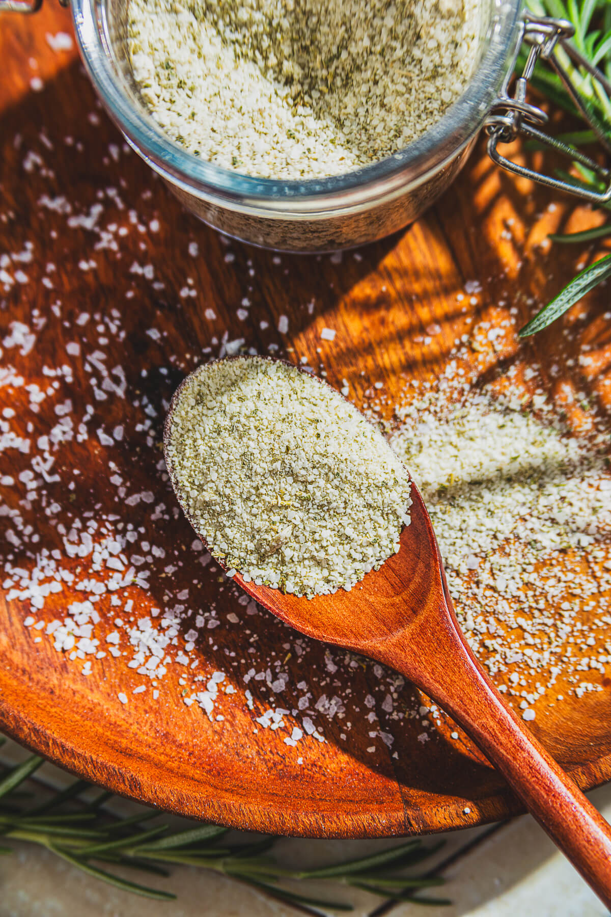 A wooden spoon of rosemary salt and a glass jar containing the same salt surrounded by fresh rosemary on a wooden platter.