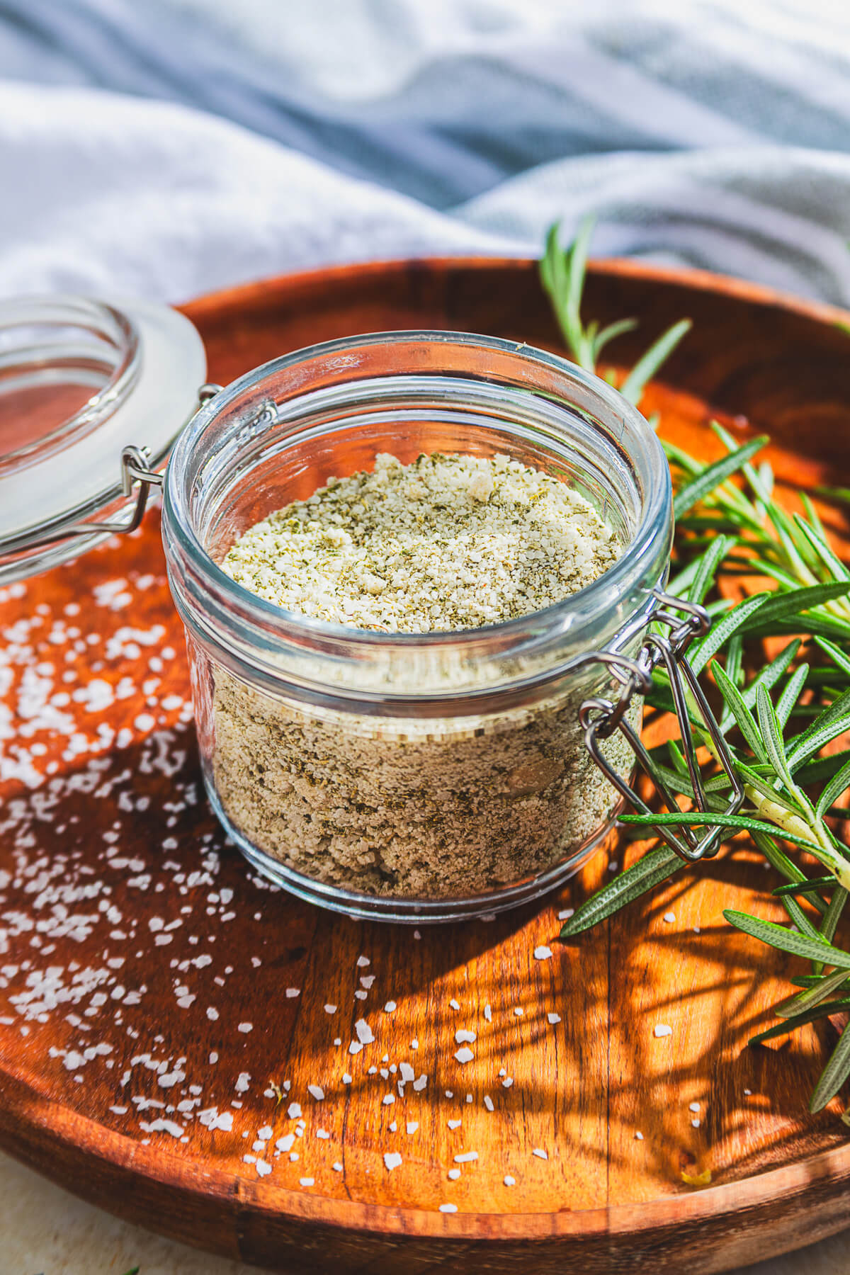 a glass jar of rosemary salt surrounded by fresh rosemary on a wooden platter.