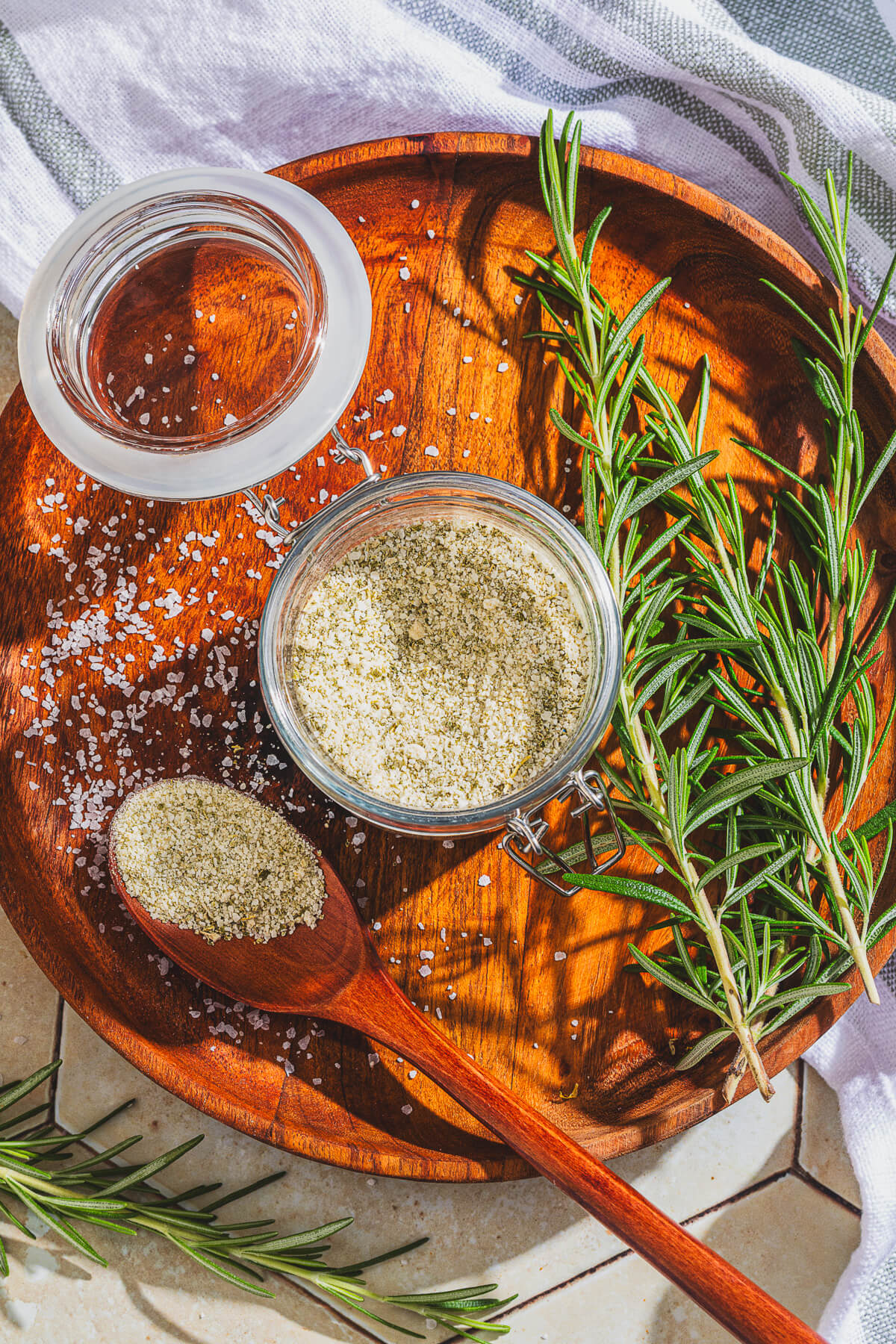 A wooden spoon of rosemary salt and a glass jar containing the same salt surrounded by fresh rosemary on a wooden platter.