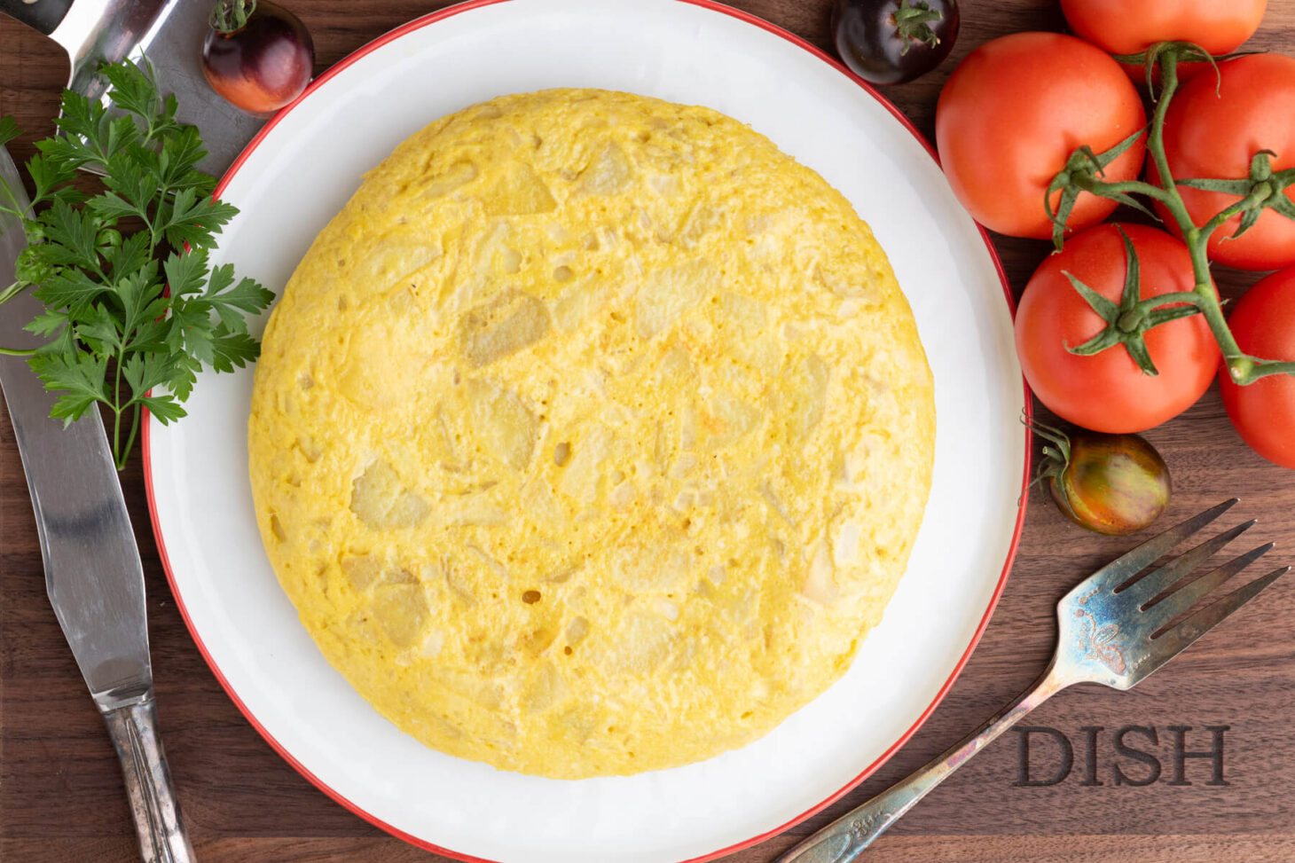 A perfect round yellow Spanish Tortilla or Tortilla de Patatas on a white plate with a red rim surrounded by tomatoes.
