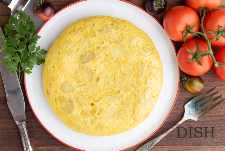A perfect round yellow Spanish Tortilla or Tortilla de Patatas on a white plate with a red rim surrounded by tomatoes.