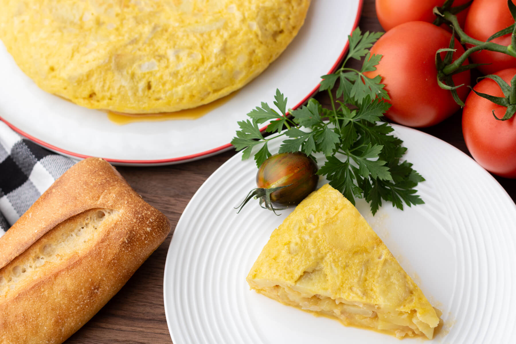 A perfect slice of yellow Spanish Tortilla or Tortilla de Patatas on a white plate surrounded by tomatoes, crusty bread, and more tortilla.
