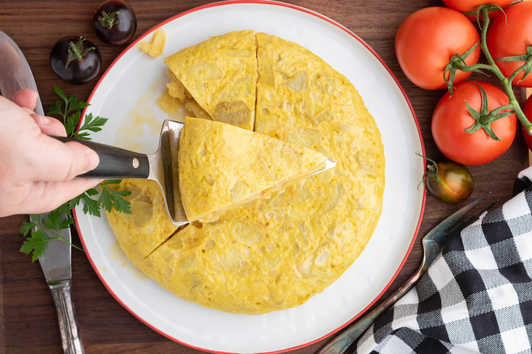 A slice being served from a perfectly round yellow Spanish Tortilla or Tortilla de Patatas on a white plate with a red rim surrounded by tomatoes.