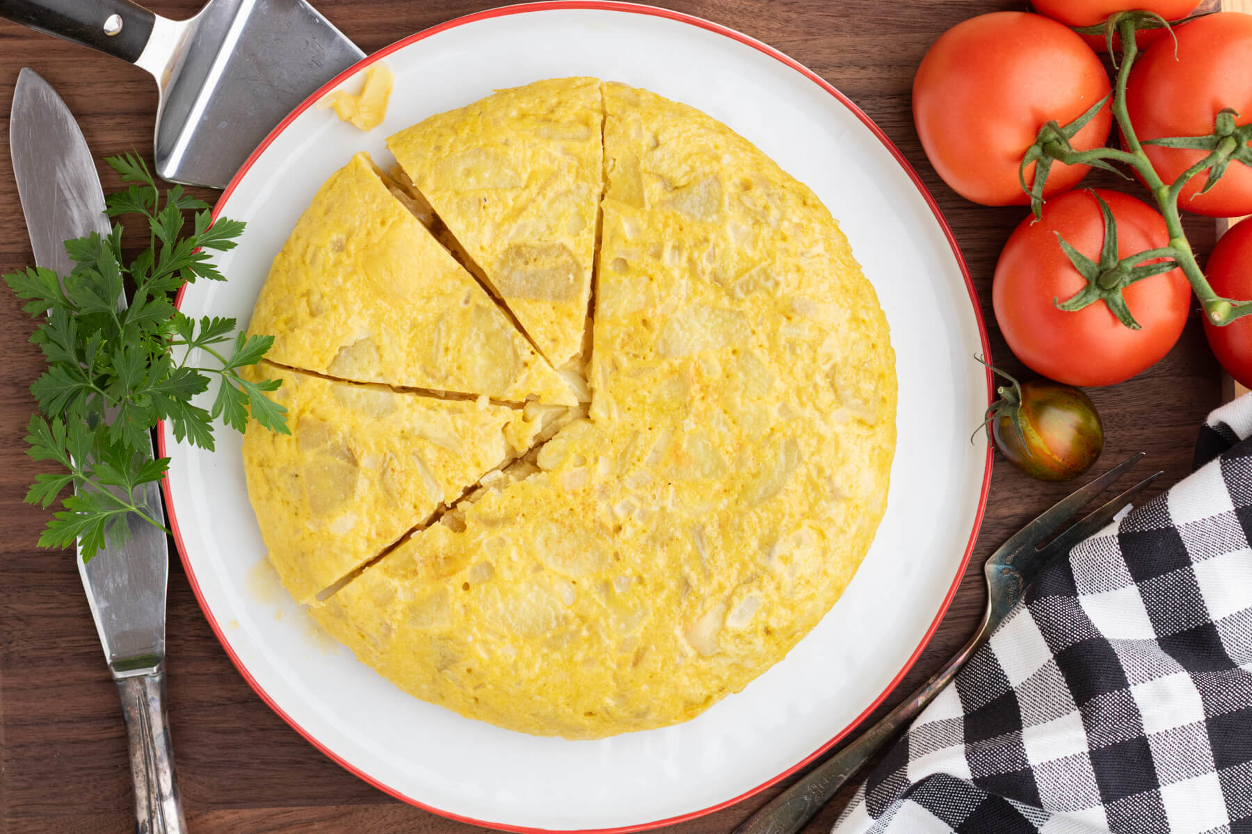 A perfect round yellow Spanish Tortilla or Tortilla de Patatas with three slices cut on a white plate with a red rim surrounded by tomatoes.