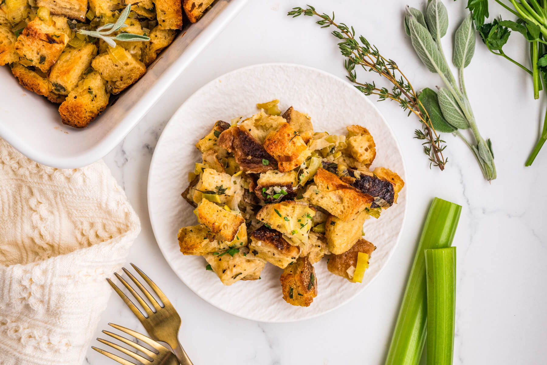 A white plate containing golden baked sourdough bread cubes seasoned with fresh herbs and aromatics on a dinner table. 
