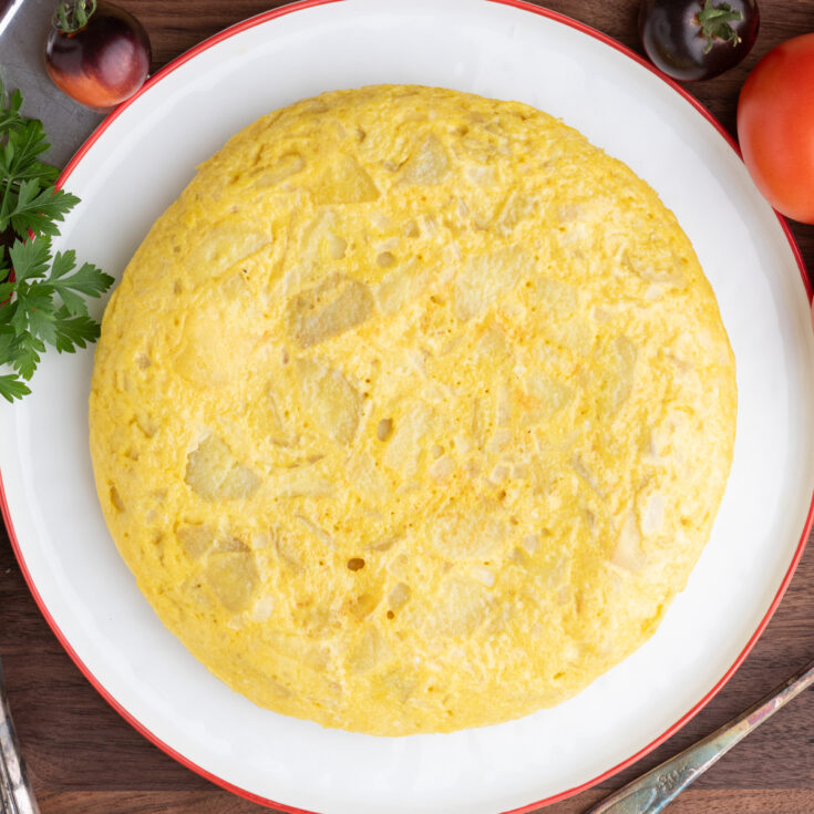 A perfect round yellow Spanish Tortilla or Tortilla de Patatas on a white plate with a red rim surrounded by tomatoes.