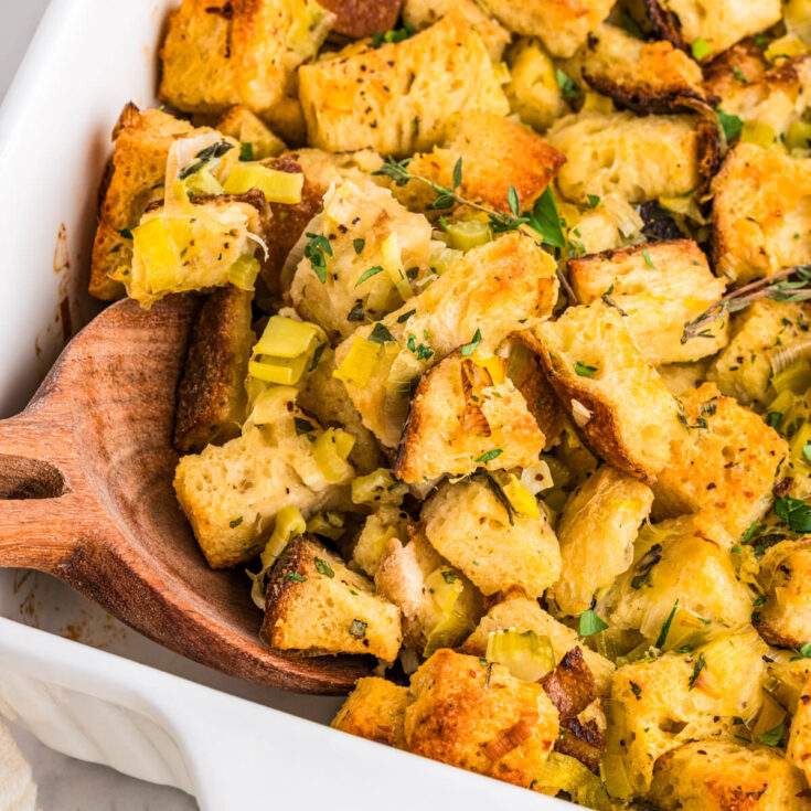 A white baking dish full of golden baked sourdough stuffing with green herbs and aromatics.